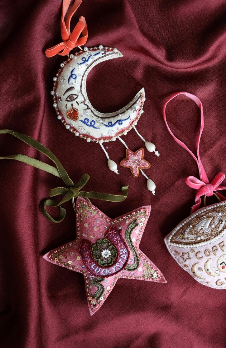 All The Luck In The World roze Lucky Star ster-ornament naast een stoffen maan-ornament op bordeauxrode achtergrond